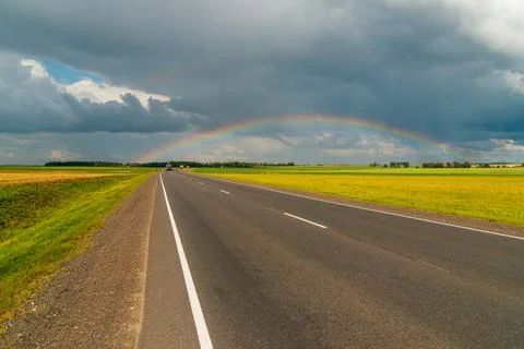 A rainbow over the road in the middle of green summer fields Stock Photos