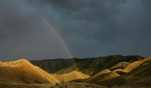 Rainbow over sunlit rolling hills beneath stormy sky Stock Photos