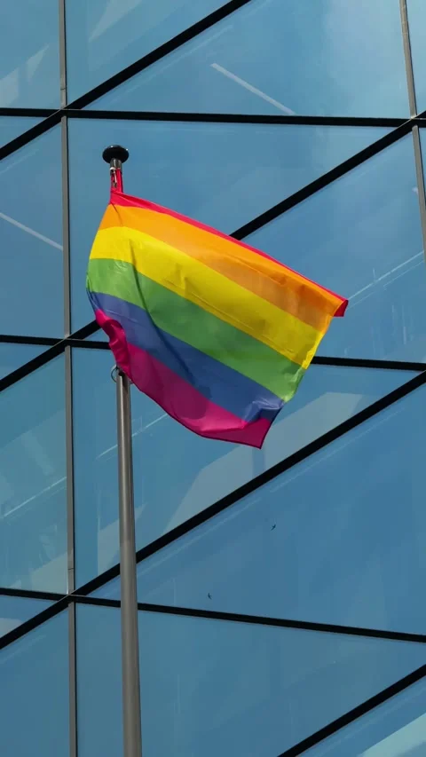 Rainbow pride flag fluttering against modern glass facade under blue sky Stock Footage 310796952
