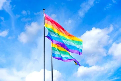 Rainbow Pride Flag Waving on Flagpole Against Blue Sky and Clouds Stock Photos