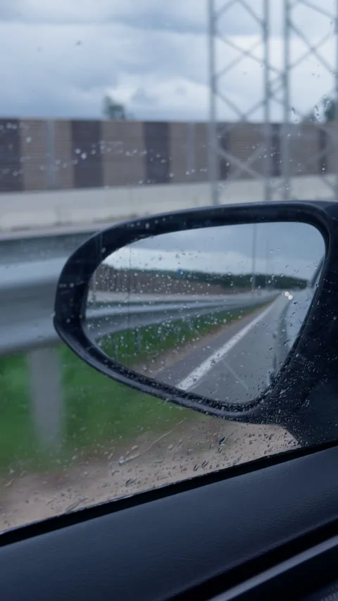 Raindrops on car side mirror reflecting wet highway beside roadside barrier on Stock Footage 345766557