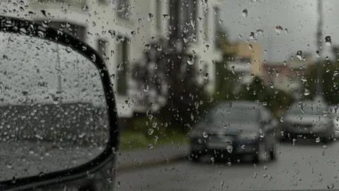 Raindrops on a car window with a blurred view of a side mirror, parked cars Stock Photos