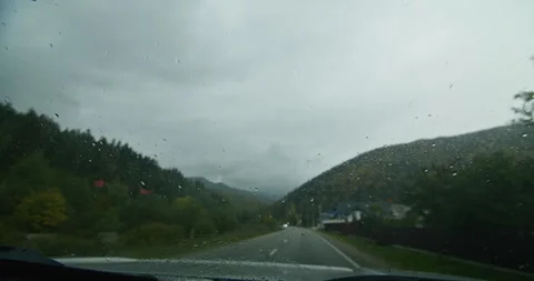 Raindrops on a car windshield with a blurred road and trees in the background Stock Footage 264998224