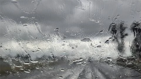 Raindrops on a car windshield obscuring the view of a cloudy sky and trees Stock Photos