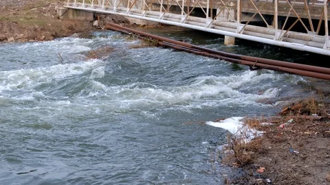 Rapid flow of river under bridge in city, rusty pipes laid across the river. Stock Footage 117700381