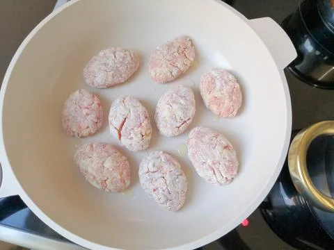 Raw breaded meat cutlets or patties arranged in a white frying pan with oil.. Stock Photos