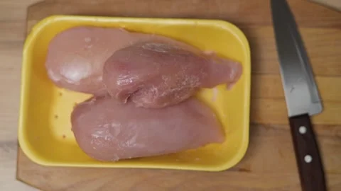 Raw chicken breasts are placed on a yellow tray while a knife rests close by on 库存影片 338298213