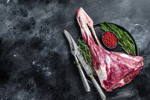 Raw lamb mutton leg meat on the bone with herbs. Black background. Top view Stock Photos