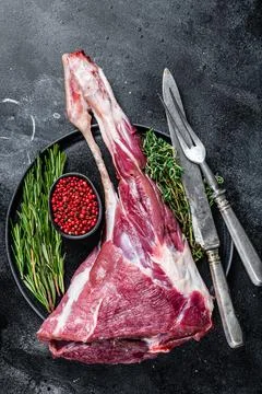 Raw lamb mutton leg meat on the bone with herbs. Black background. Top view Stock Photos