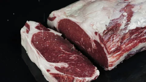 Raw marbled ribeye beef steak sprinkled with salt on black tabletop Stock Footage 169720536