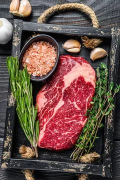 Raw prime rib eye beef meat steak in a butcher wooden tray with herbs. Black Stock Photos