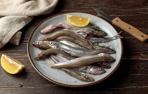 Raw small fish on a plate with a slice of lemon. Close-up on a dark wooden Stock Photos