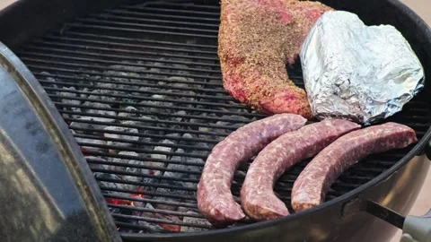 Raw tri-tip beef and sausages sit on a charcoal grill grate Stock Footage 345536688