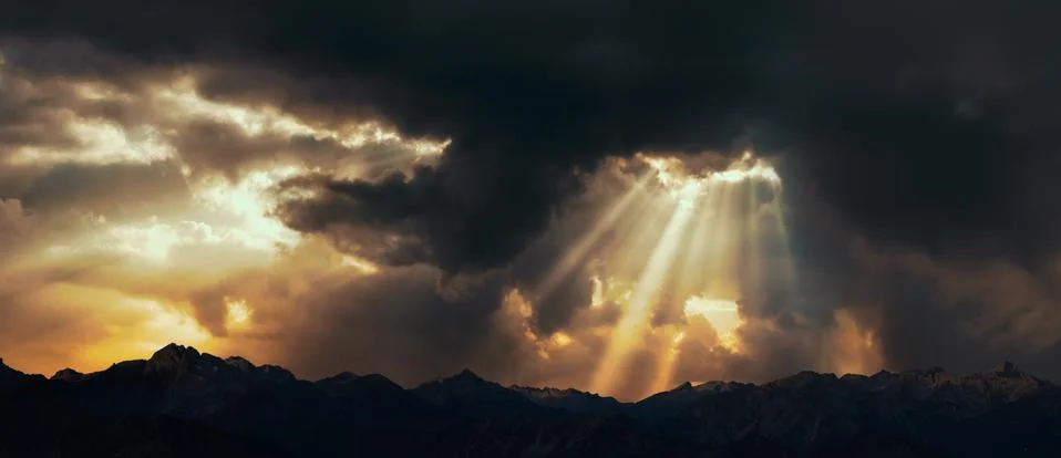 Rays of light shining through dark clouds over mountains. Cinematic scene. Stock Photos