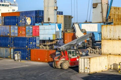 Reach stackers and cranes move containers near bulk carrier on sunny day Stock Photos