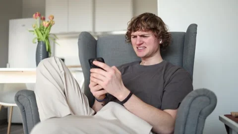 Reacting with frustration while looking at his smartphone, a young man sits in a Stock Footage 302633982