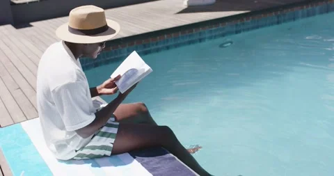 Reader is holding book on striped towel by pool, is dipping feet after pausing Stock Footage 335625708