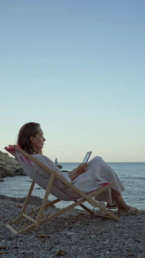 Reading on the beach while sitting in a chair by the water in the evening Stock Footage 342788046
