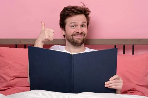 Reading an exciting book sitting on bed at home showing thumb up Stock Photos