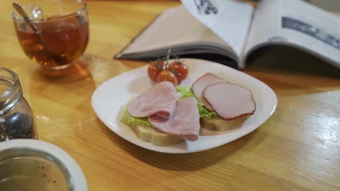 Reading magazine during breakfast in kitchen. Ham sandwich with tomatoes on Stock Footage 96194965