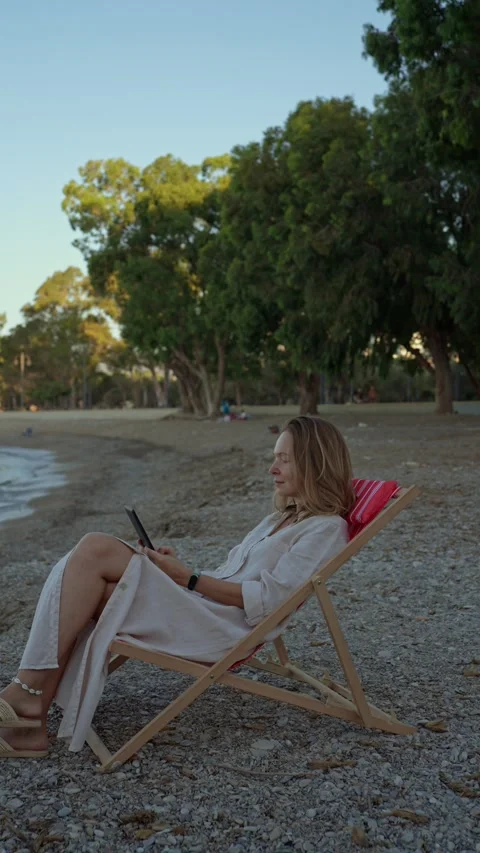 Reading by the water on a beach in the evening with trees in the background Stock Footage 346016808