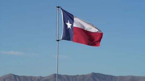 Real time shot of a Texas flag slowly waving in blue sky above desert hills Stock Footage 166876520