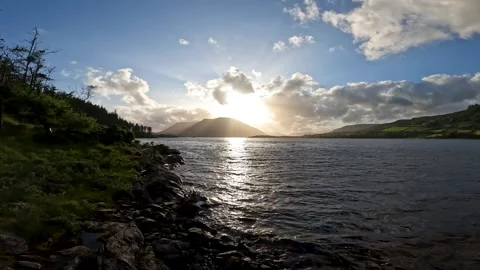 Real time sunset over mountain watching from the shore line of a river lapping Stock Footage 309973682