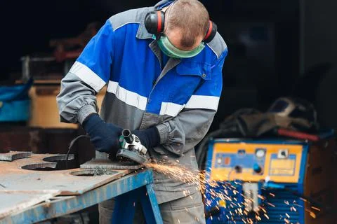 Real worker in working clothes grinds metal surface and sparks fly. person at Stock Photos