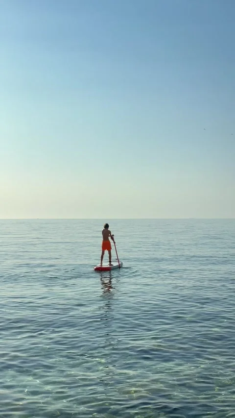 Rear view of a boy stand-up paddleboarding on a calm, transparent sea under.. Stock Footage 338444033