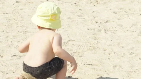 Rear view close-up of toddler boy in bucket hat playing in sand on beach Stock Footage 330542193