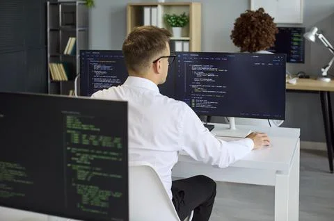 Rear view of developer coding at dual monitors in contemporary tech office Stock Photos