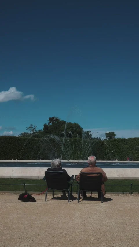 Rear view of an elderly couple sitting on chairs beside a fountain with a small Stock Footage 335382385