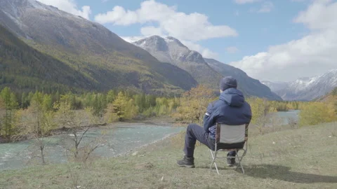 Rear view of guy sitting on a riverside in mountans valley and contemplating 4k Stock Footage 85495141