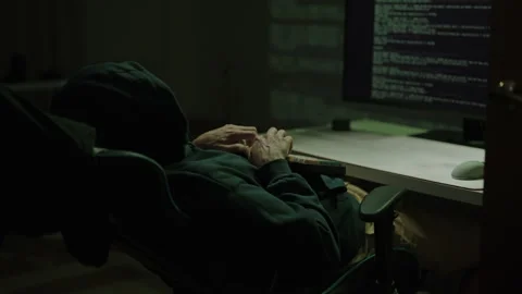 Rear view of hooded hacker typing code on keyboard in dark room, working on Stock Footage 323470440