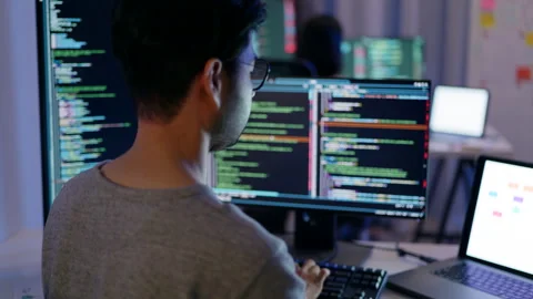 Rear view of a male programmer working on multiple monitors filled with code. Video stock 310589111