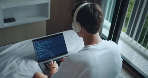 Rear view man freelancer programmer in headphones sitting on bed in bedroom and Stock Footage 251023397