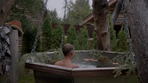 Rear view of man immersing himself in huge tub of hot water infused with forest Stock Footage 221940575