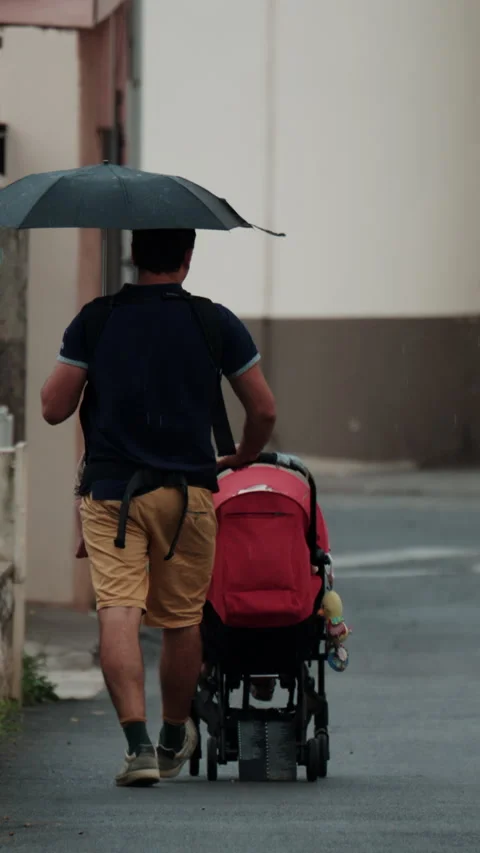Rear view of a man pushing a stroller while holding an umbrella on a rainy urban Stock Footage 339742225