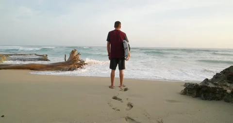 Rear view. Man with surfboard standing on tropical beach against Atlantic Ocean. Stock Footage 182078726