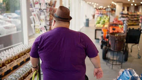 Rear view: An overweight man in a purple T-shirt and a brown hat walks through Stock Footage 252183362