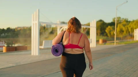 Rear view of a plus size female holding a fitness mat walking in the morning. Stock Footage 344248209