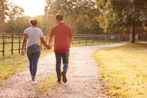 Rear View Of Romantic Couple Walking Hand In Hand Along Country Lane At Sunset Stock Photos