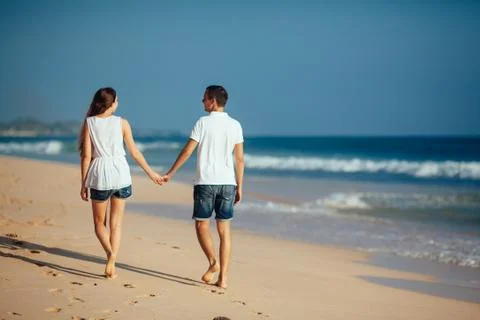 Rear view of romantic happy couple walking on beach holding hands at blue sky Stock Photos