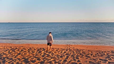 Rear view of young man in baseball cap and hoodie approaching the sea on beach Stock Footage 185773313