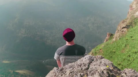 Rear view of a young man in a hat, sitting on a stone with his back against the Stock Footage 185791619
