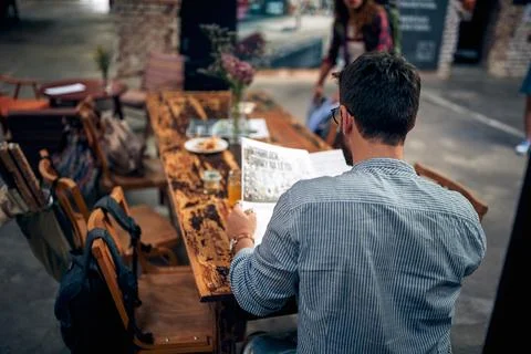 Rear view of young man reading newspaper in cafe, sitting alone at table. Stock Photos