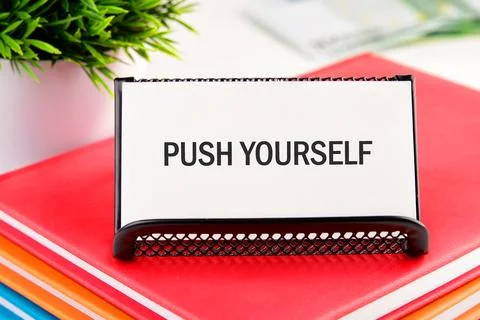 A rectangular white sign with the words PUSH YOURSELF rests on colorful books. A Stockfoto's