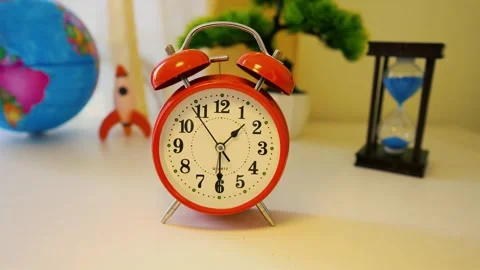 Red alarm clock on a desk with a globe, rocket, and hourglass in the backgrou Stock Footage 317736793