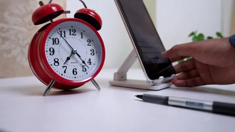 Red alarm clock next to a smartphone on a stand, with a hand typing Stock Footage 317131671