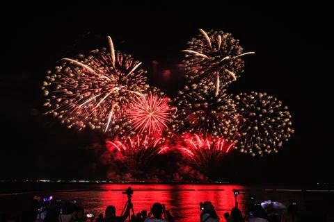 Red And Gold Fireworks Exploding Over Water With Silhouette Crowd On Shorel.. 库存照片